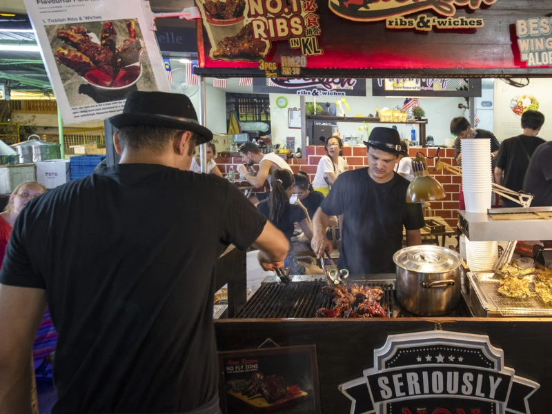 Sizzling BBQ pork ribs grilling at a popular Jalan Alor street food stall, a mouth-watering culinary experience safely and effortlessly enjoyed on your Kuala Lumpur day trip private car tour.