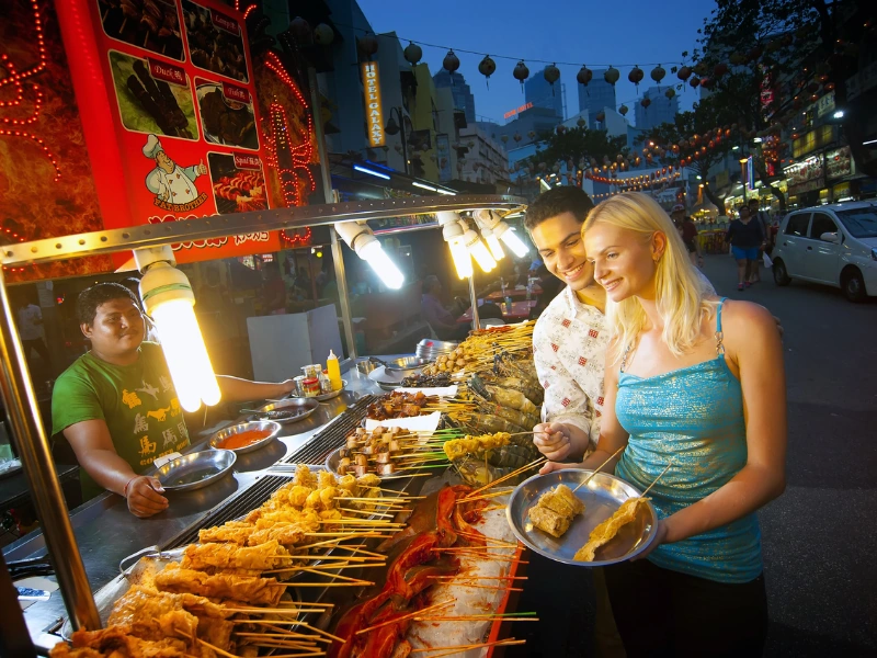 Happy tourists choosing delicious local skewers at a brightly lit Jalan Alor street food stall, an authentic culinary adventure made easy with your Kuala Lumpur day trip private car service.